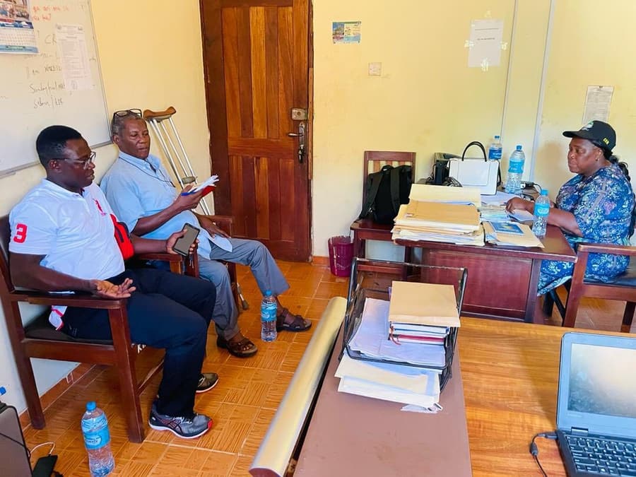 Board members meeting WESOFOD staff around a desk at the head office, a pair of crutches resting against one of the chairs.