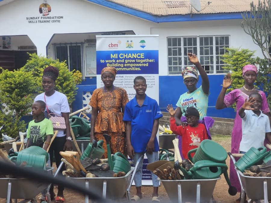 Adults and children stand with wheelbarrows of watering cans and hand tools in front of an MI-Chance banner at the Sunday Foundation Skills Training Centre.