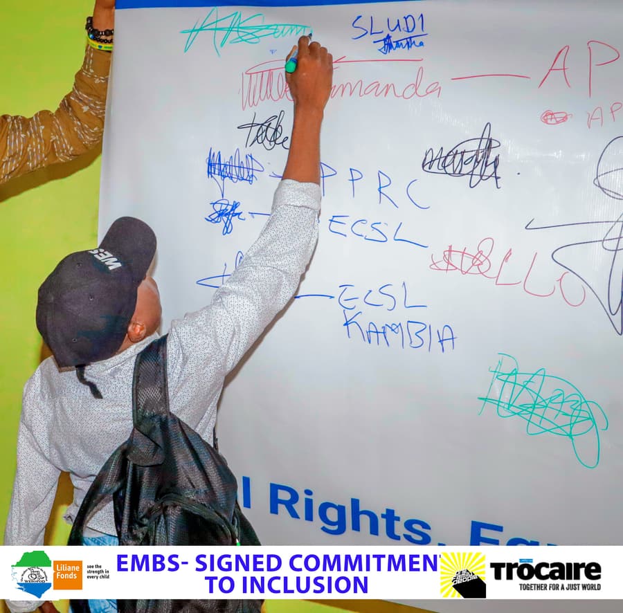 A hand signing a large commitment poster in coloured marker, with signatures from SLUDI and political party representatives already on it.