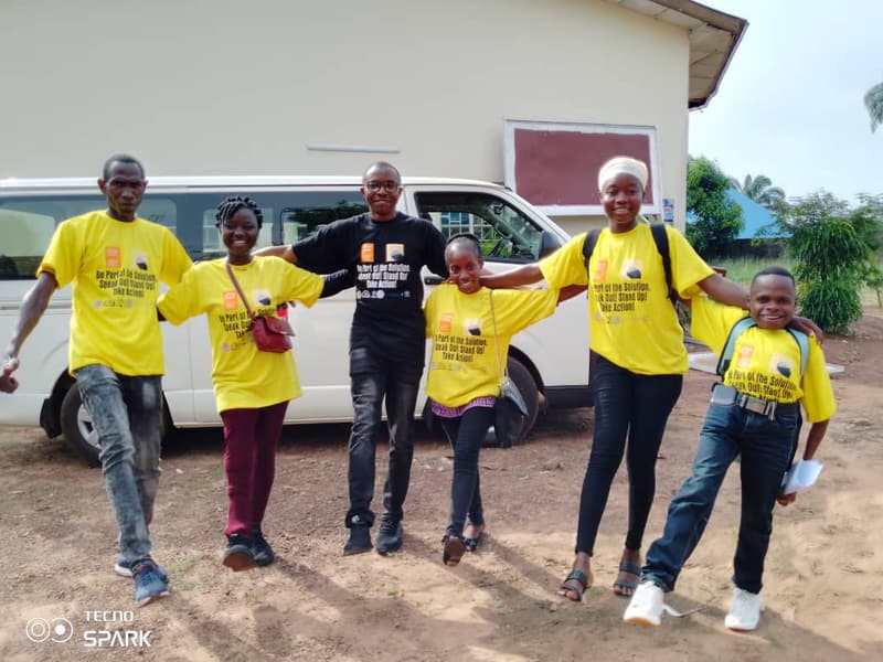 Six young people in yellow campaign T-shirts link arms and step forward together in front of a minibus