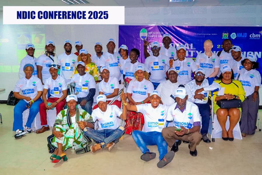 Conference participants in white caps and T-shirts, several of them with crutches or mobility aids, pose together at NDIC 2025.