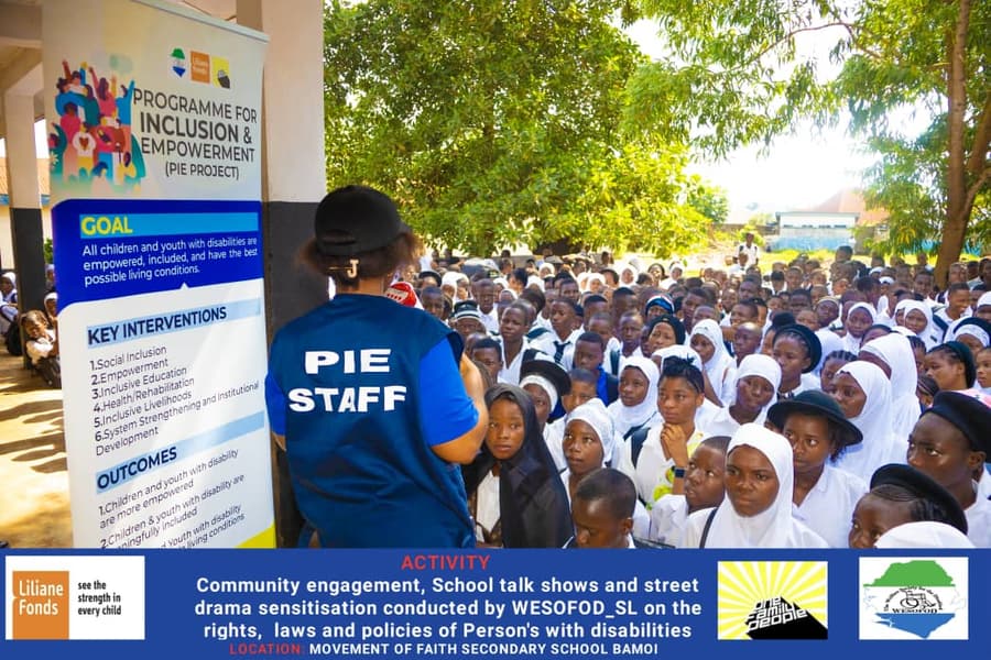 A WESOFOD-SL staff member in a PIE Staff vest addresses a large crowd of secondary-school students beside the programme banner.