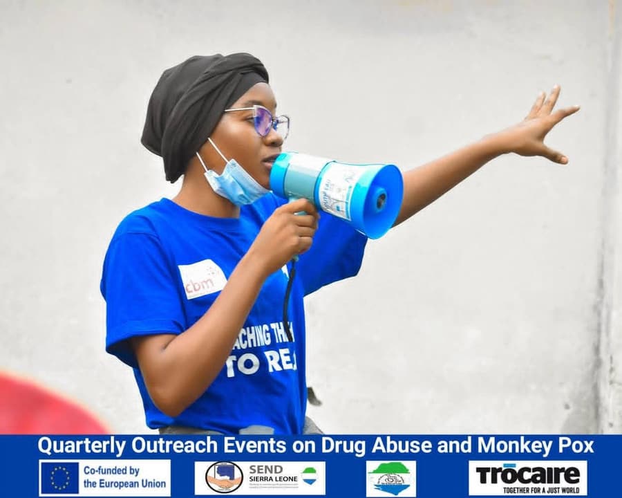 A young woman in a WESOFOD-SL T-shirt speaks through a megaphone, one arm stretched out, at a quarterly outreach event on drug abuse and mpox.