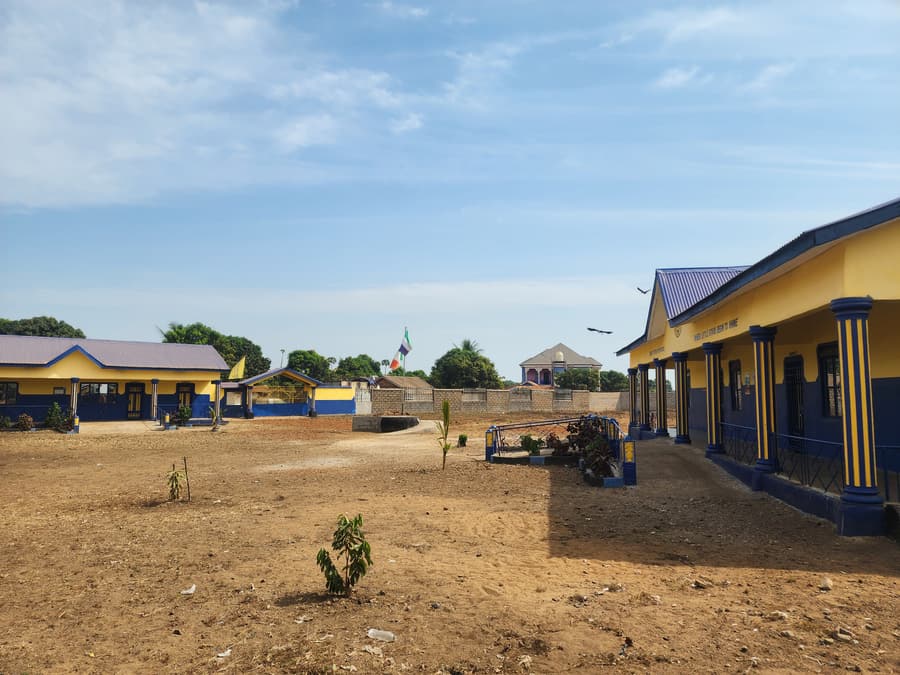 Blue and yellow buildings of the Village of Hope in Kambia around an open sandy yard with young trees